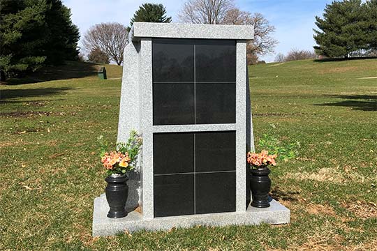Cremation columbarium in gray granite with eight cremation niches with black granite fronts and two black vases on each side.